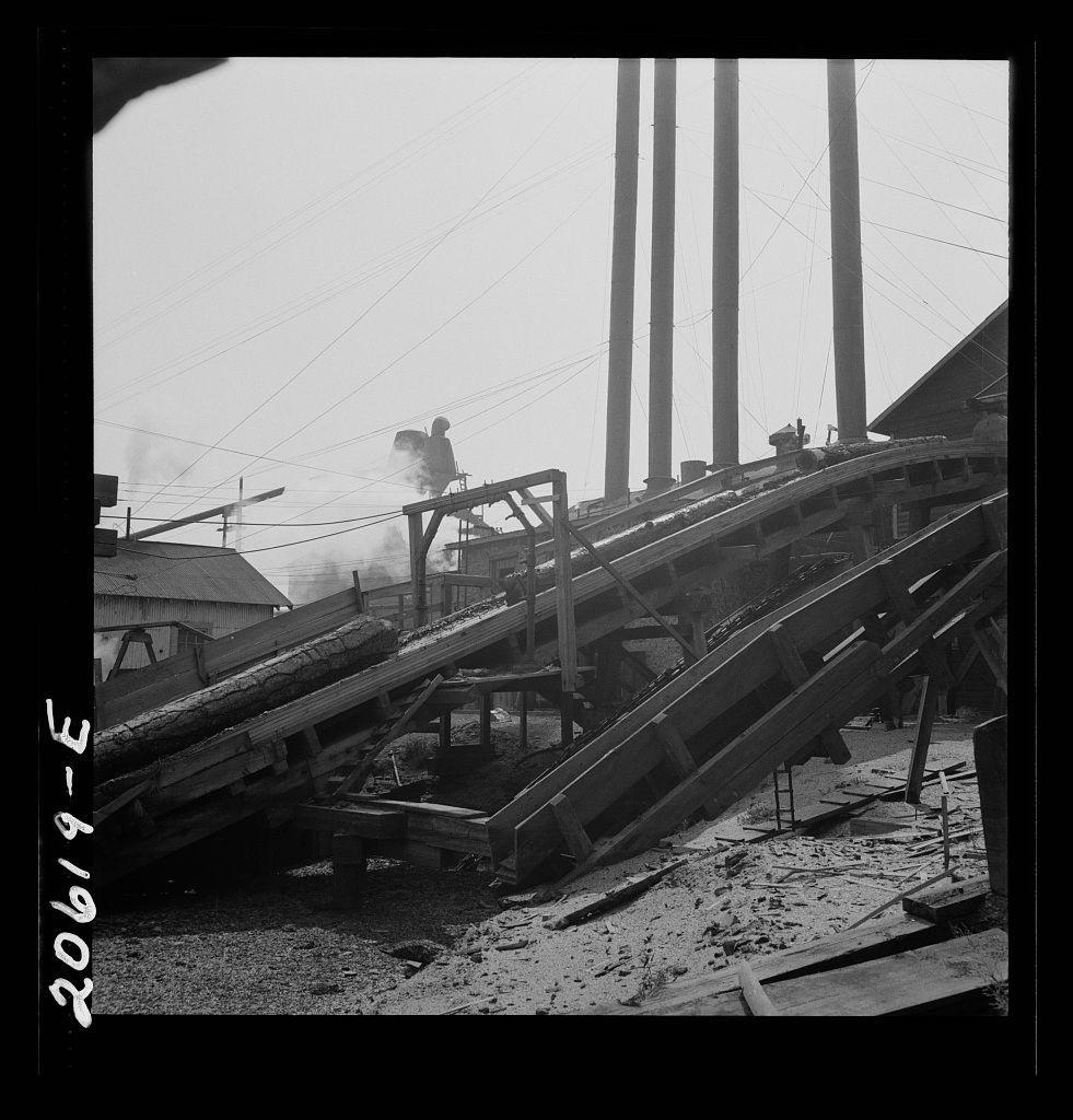 The image depicts a scene from what appears to be an industrial lumber mill. The photograph is in black and white, giving it a vintage feel that suggests historical significance. It captures the essence of laborious manual work.

In the foreground, there's a prominent wood chute or conveyor belt system extending diagonally across the frame. It has multiple levels with logs on top, indicating its function for transporting timber from one point to another within the mill complex. The structure seems sturdy but shows signs of wear and disrepair in some areas where supports appear warped.

A worker is visible atop a higher level of this chute, seemingly engaged in operating or supervising the movement of wood. Their posture suggests exertion, likely indicative of physically demanding labor typical for someone working at such mills during that era.

The environment around includes various industrial elements like metal poles and infrastructure supporting electricity transmission lines, which are prominent above. These structures add to the overall impression of an active heavy industry operation.

In the background, there is a suggestion of smoke or steam rising from behind some buildings, possibly indicating ongoing processing activity within other parts of the mill complex. The sky appears overcast with diffuse lighting conditions, adding to the somber tone of industrial lab [...]