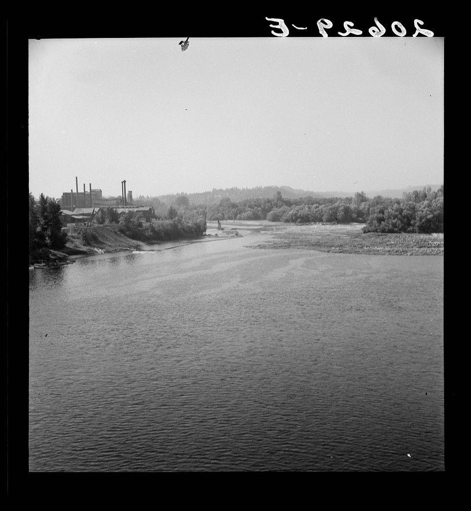 The image is a black-and-white photograph depicting an industrial landscape. In the foreground, there's calm water that reflects some of the sky and surrounding scenery. Further back on the left side, we see what appears to be a factory or power plant with large structures such as chimneys or smokestacks rising above trees and other vegetation. The land around this facility is flat and extends into the distance where more industrial buildings are faintly visible against a horizon lined by dense woodland.

In the top right-hand corner of the image, there's handwritten text in what seems to be Cyrillic script that reads "3-PSOS". This could indicate either a date or an identifier related to the photograph. The overall composition suggests this might have been taken during a time when industrialization was prominent and likely serves as historical documentation.

This photo does not display any explicit human subjects, hence there are no details regarding gender or race of individuals in motion within it.