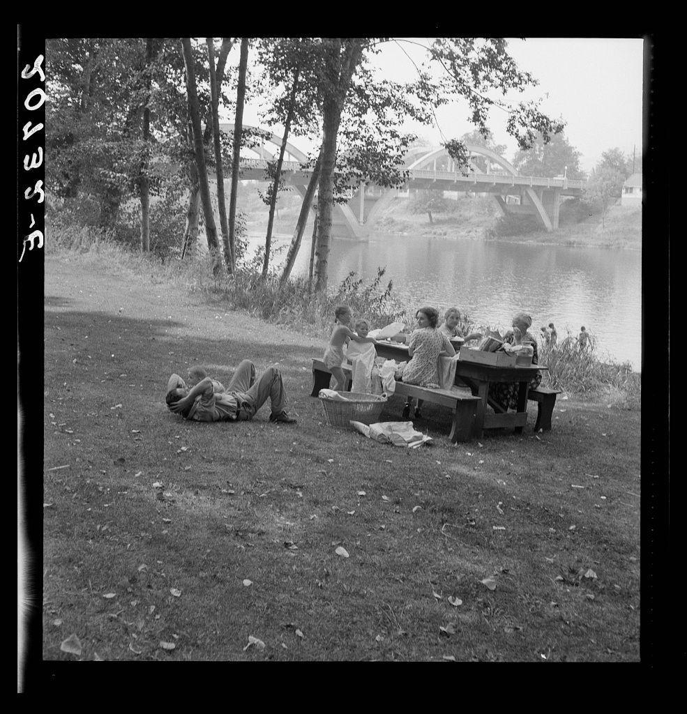 The image is a black-and-white photograph depicting several people enjoying an outdoor picnic by the riverside. There are groups of individuals sitting at tables, some engaged in conversation and others focused on reading materials like newspapers or books. One person can be seen lying down next to one such table with their head resting against it as if they were taking a nap or relaxing.

In the background, there is a bridge spanning across what appears to be a river or lake, surrounded by lush greenery and trees. The scene suggests a leisurely day spent in nature during an outdoor family gathering. Notably, this photograph seems to capture a moment of relaxation among individuals from different generations gathered for a Sunday picnic near the Rogue River, as mentioned in Oregon's Josephine County Grants Pass context provided.

The image conveys a sense of community and shared enjoyment while highlighting the simple pleasures of spending time outdoors with loved ones on what appears to be an enjoyable sunny day.