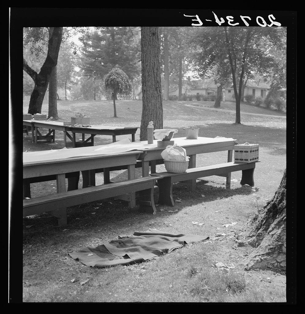 The image is a black and white photograph depicting an outdoor setting that appears to be from the early 20th century, based on its monochrome quality. In this scene, there's a picnic area with wooden benches surrounding a long table covered in what seems like newspapers or a makeshift tablecloth. On one side of the table, various objects are placed including containers possibly containing food and utensils, as well as baskets that likely held other items for the outing.

The ground is grassy with patches visible where soil has been exposed, suggesting frequent use at this site. There's also evidence of wear on a large piece of fabric spread out in front of one bench, perhaps used earlier or forgotten during cleanup after the picnic concluded. In the background stands tall trees and houses that blend into their wooded environment.

The word "3-beros" is written across the top right corner, likely indicating either an informal caption or part of a larger message not fully visible within this frame. The overall impression conveyed by the image is one of communal gathering during warmer weather in what seems to be a small-town park setting, evoking memories and simplicity associated with early American rural life.

Further context about the photograph suggests it was taken on "California Day" at Grants Pass, Josephine County, Oregon as indicated by an additional caption. Thi [...]