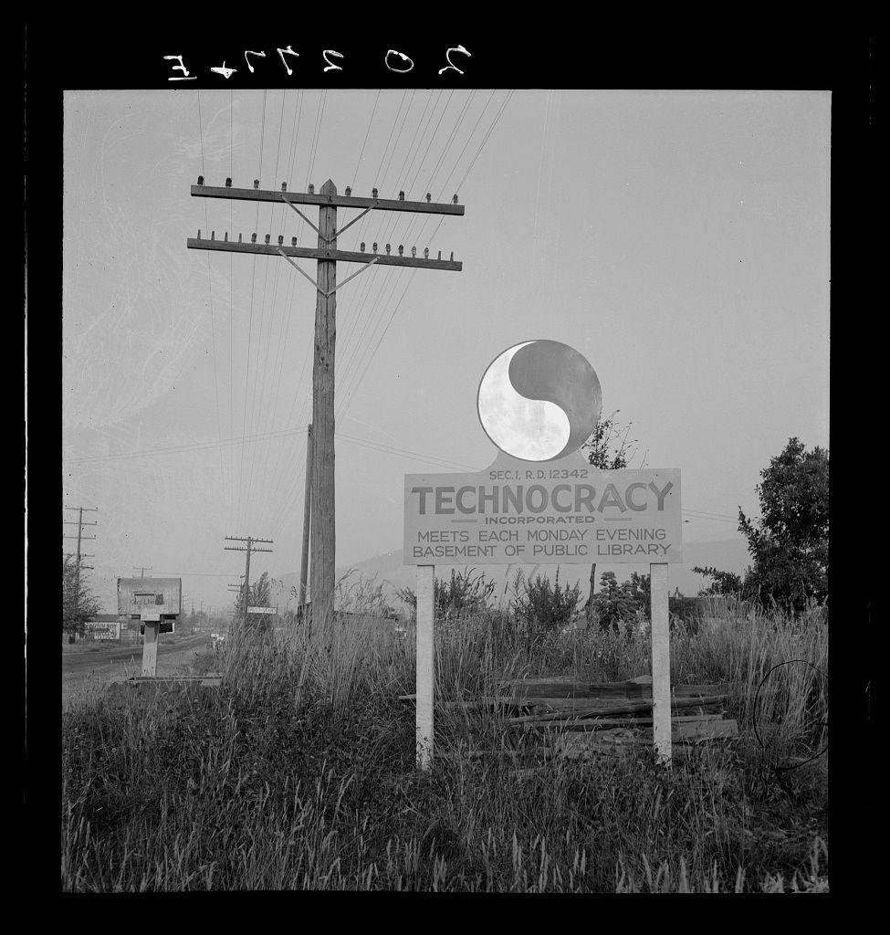 The image depicts a black and white photograph featuring an old power pole with numerous wooden crossbars against the sky. In front of this setting, there is a signpost that reads "TECHNOCRACY" in bold capital letters at its center, accompanied by smaller text underneath indicating it's associated with SEC.R.D.I 1234-0 and includes mentions about public library meetings held on Monday evenings. Behind the power pole stands a large circular emblem depicting what appears to be an eye or celestial body within another shape that resembles a circle divided into four quadrants, suggesting a symbolic representation of technology or surveillance. The surrounding area is overgrown with tall grasses, hinting at rural or neglected urban surroundings.