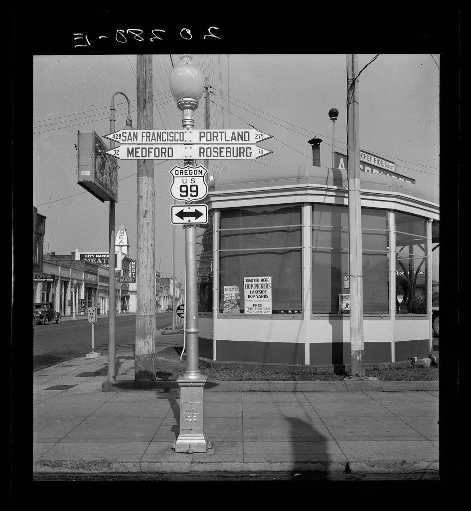 The image is a black and white photograph depicting an intersection with multiple directional street signs. The most prominent sign indicates the distances to various cities such as San Francisco, Portland, Medford, Roseburg in Oregon, along U.S. Highway 99. Other smaller signs are visible for local businesses like "Chef Eide House" restaurant, a health food store called "Health Mart," and an advertisement for a "Hop Yard." The photograph is taken during daylight with clear skies, showcasing the urban environment of that era.