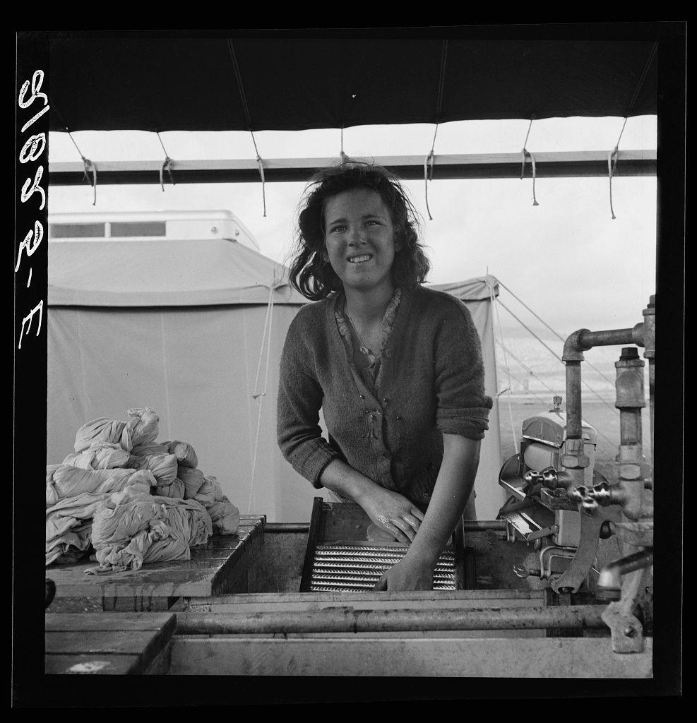 The image is a black and white photograph depicting an individual working with laundry in what appears to be a mobile or temporary setup, possibly related to migrant labor. The person stands beside a washing machine loaded with clothes. Behind them are bags of clothing stacked on top of each other. A large tent-like structure provides shelter for the area where the work is being done. Visible equipment includes pipes and valves associated with water supply systems, suggesting that this might be part of a temporary camp setup providing facilities like laundry services.

Additional context provided in the caption mentions Merrill FSA (Farm Security Administration) camps specifically located in Klamath County, Oregon during certain periods when such facilities were active for accommodating migrant workers. The image is labeled as being from Young migrant girl makes use of facilities provided for cleanliness and associated with a specific archival reference or photograph number: "6495." This suggests that the photo was taken by Dorothea Lange, who documented various aspects of American life during her work under the Farm Security Administration's photographic program.