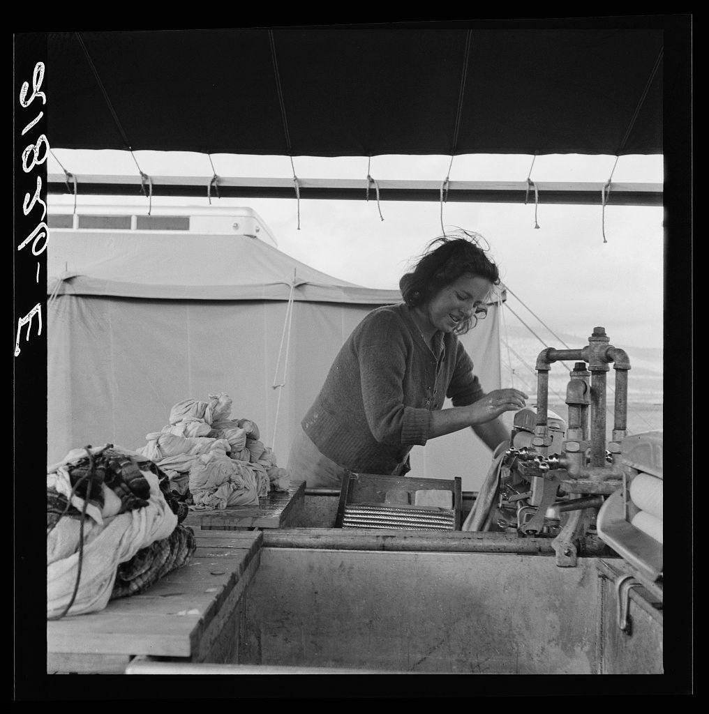The image is a black and white photograph depicting an individual interacting with some machinery. The person appears to be working on or operating the equipment, which has various pipes and valves attached to it. They are wearing glasses, a sweater, and appear focused on their task.

In the background, there's a large tent-like structure that suggests this scene may take place in a temporary setup such as campsite or an outdoor facility. There is also some laundry visible near what seems like sleeping quarters with rolled blankets stacked up outside of it.

The photograph has text written across the top and bottom left corners which might indicate context about its origin, such as "FSA" (Federal Security Agency) and references to a migrant girl using facilities provided for cleanliness in Merrill FSA camp. The specific location is mentioned as Klamath County, Oregon. There's also an additional reference indicating this may be related to general caption 62.

The image has a historical feel due to its monochrome nature and the setting it portrays which suggests that it might have been taken during or around World War II era when such camps were used for housing displaced persons.