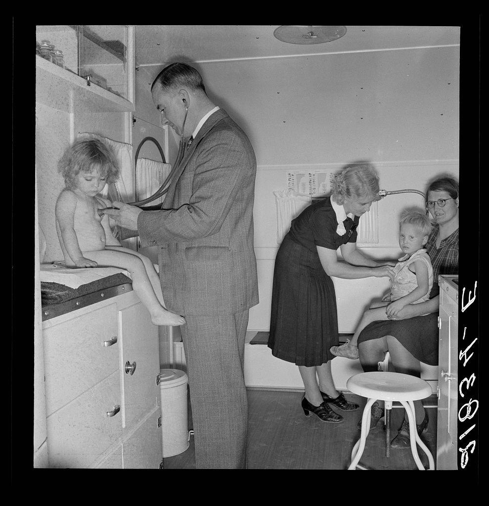 The image is a black and white photograph depicting an indoor medical examination scene. In the foreground, a man in professional attire—a suit with a tie—is using a stethoscope to examine a young child sitting on a high stool or cabinet next to him. The examined child appears barefoot and has curly hair.

Behind this interaction, two women are attending to another child seated on their laps. One woman is wearing glasses, while the other's face isn't fully visible due to her positioning in the background. They both seem engaged with the children they're caring for or assisting.

The room contains various objects such as a small table holding what appears to be medical supplies and a cabinet with drawers that likely contain more equipment. The setting looks like an improvised clinic, possibly within a trailer given its compact nature and functional design without ornate details.

In terms of context provided by the caption "Doctor examining children in trailer clinic," it suggests this photograph is taken at a mobile camp run by the Farm Security Administration (FSA), which was part of efforts to support rural communities during the Great Depression. The specific location mentioned, Klamath County, Oregon, indicates that these clinics were likely set up temporarily for medical services where they weren't readily available.

The overall atmosphere suggests an era when such  [...]