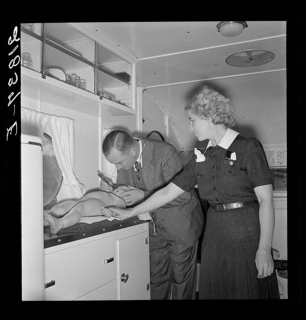 A black and white photograph depicts a medical examination taking place. A man, presumably a doctor or nurse due to his attire of rolled-up sleeves and pocket protector with pens, is attending to an infant on the operating table within what appears to be a makeshift clinic set up in a trailer home. He uses tools like scissors near the child's legs while another person observes from beside him; this individual seems to wear a service uniform indicating she could possibly be part of medical or administrative staff at that time and place, suggesting an era where women were not yet commonly seen as frontline healthcare providers.

The setting is utilitarian with cabinetry for storage. In the background are shelves containing jars, bowls and plates atop glass cabinets which further implies this scene takes place in a temporary structure designed to serve health services amidst mobility or homelessness circumstances like those associated with migrant labor during certain periods of American history. The image conveys a sense of urgency yet also care as there is no visible distress from the subjects despite an apparent medical examination underway.

Additional information suggests that this photo may have been taken at Merrill, possibly referring to Camp Merril which was part of the larger network of camps housing migrant laborers during times when such groups were not always we [...]