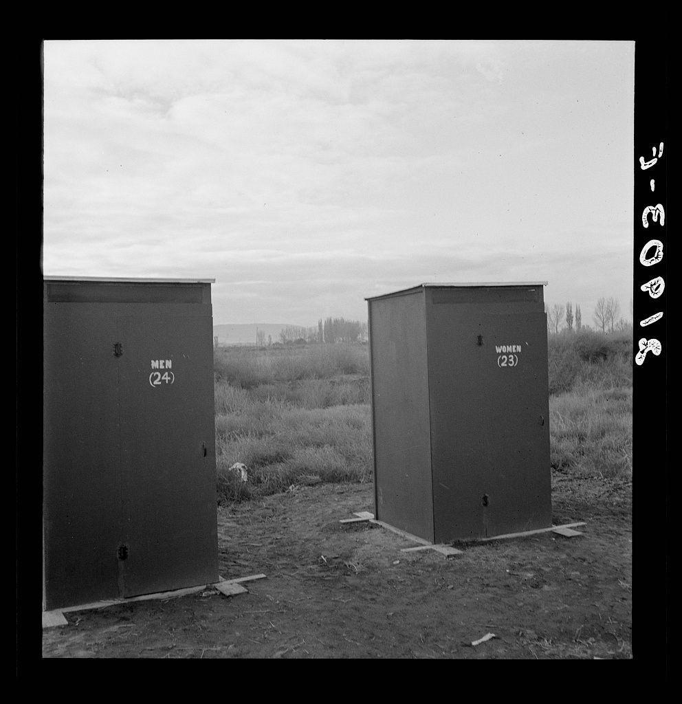 The image displays two portable toilets situated in a rural outdoor setting. These are labeled as "MEN (24)" and "WOMEN (23)," indicating the number of available facilities for each gender, suggesting that they may be located within or near an encampment such as a farm labor camp during a time when temporary housing was required by migrant workers. The black-and-white composition adds to the historical feel of the image, with barren trees and grass in the background under a cloudy sky enhancing this impression.

Additional context about these portable toilets is available from Twenty-four portable toilets, mobile camp (FSA - Farm Security Administration) equipment located at Merrill, Klamath County, Oregon. This reference suggests that the photo was taken as part of documentation by the FSA during its efforts to support agricultural workers and their living conditions in rural areas such as those found within or near farms.

The photograph captures a significant aspect of American history where migrant laborers lived temporarily while working on seasonal farm jobs. The equipment used for housing these individuals, including portable toilets like shown here, underscores the challenges faced by them during that period concerning basic amenities and infrastructure to support their living conditions away from home.