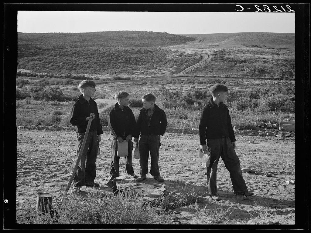 The image depicts a group of four individuals, who appear to be boys or young men, standing in an outdoor rural setting. The landscape behind them is hilly and barren with sparse vegetation under clear skies.

Each individual holds different objects; from left to right: the first person has what seems like a shovel upright on the ground beside him, another two have paper bags possibly containing items for examination, while the last one appears empty-handed but stands apart slightly. All are dressed in dark clothing and casual footwear suitable for outdoor work or activity.

The photograph is monochromatic (black-and-white), adding to its historical ambiance as it seems older than current times given the attire of individuals and overall quality of photo which suggests an era from past decades, possibly mid-20th century judging by photographic style. The setting appears remote with no immediate signs of modern infrastructure or habitation.

There's a note "O.S.Rolls" handwritten on the top right corner in what looks like cursive handwriting that might be indicative of photographer’s watermark or signature.