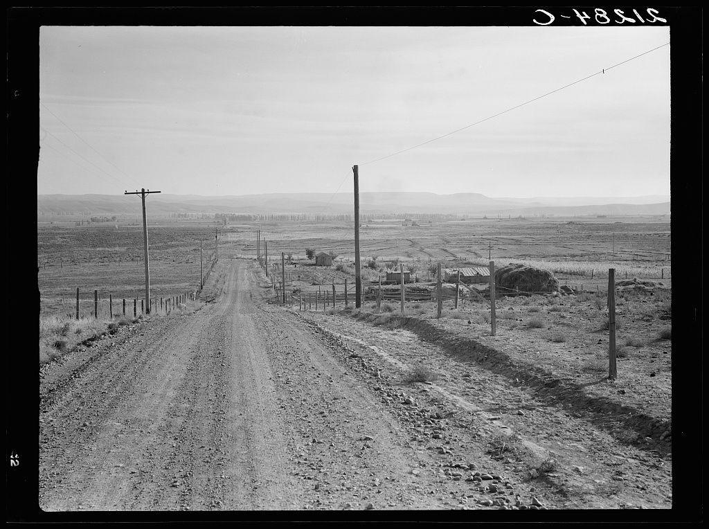 The image is a black and white photograph depicting a rural landscape, likely from the early to mid-20th century. A dirt road stretches out into the distance with fences lining its sides on both ends of the frame. Sparse vegetation dots the otherwise barren land which extends towards rolling hills in the background under an expansive sky. Several structures can be seen, including what appears to be a dilapidated barn and some smaller sheds or houses, suggesting this might be farmland or ranchland.

The photograph is marked with "D-14815" at the top right corner, indicating it may have been catalogued as part of an archive. The text on the bottom left indicates that additional information can be found in Owyhee project landscape and references a specific number within this context (General caption number 66). This suggests the photograph is from a larger collection or study related to landscapes around Vale, Malheur County, Oregon.

The overall impression of the image conveys a sense of isolation and simplicity associated with rural life during that era.