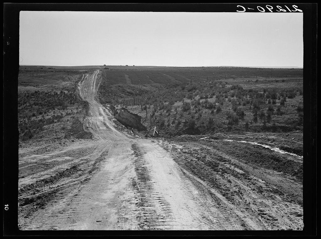 The image depicts a black and white photograph of an unpaved, dirt road cutting through a vast rural landscape. The road appears to be deeply rutted with tire tracks crisscrossing the surface, suggesting frequent use despite its rough condition. On either side of the road are barren fields or dry brushland stretching into the distance where sparse vegetation can be seen scattered across the plains.

The sky above is clear and devoid of clouds, hinting at a bright but perhaps overcast day due to the lack of distinct shadows on the ground. The terrain appears uneven with slight undulations in elevation visible as gentle hills or mounds rise from either side of the road. There are no signs of water bodies like rivers or lakes within the immediate vicinity.

In the upper right corner, there's a handwritten label "J-OPSLA," which could be an acronym for a location, project, or individual related to this photograph. In the lower left corner is another mark indicating the image number as 103942.

Additional context provided by the caption states that this road was constructed by the Civilian Conservation Corps (CCC), part of President Franklin D. Roosevelt's New Deal program during the Great Depression, which aimed at relief and creation for unemployed Americans through various public works projects including roads building across America.

The image is titled "Landscape on top  [...]