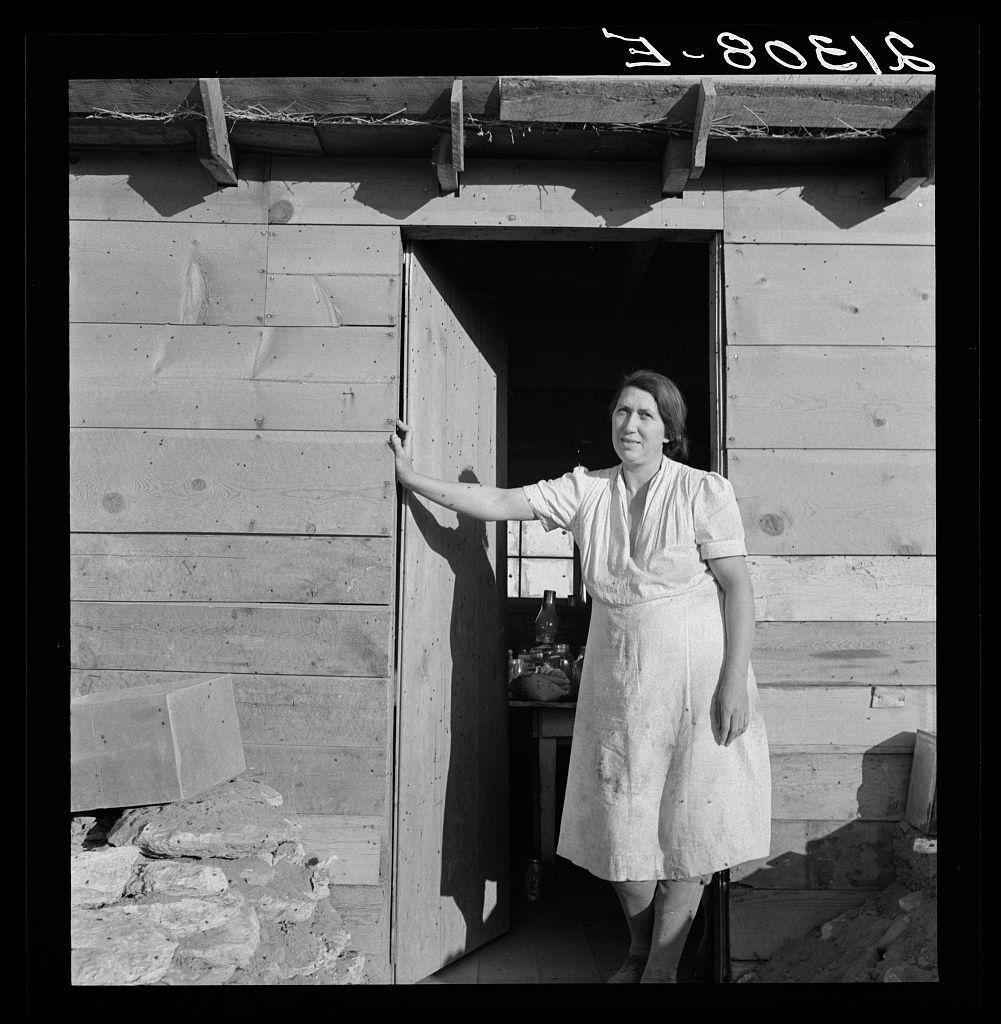 The image is a black and white photograph depicting an older woman standing in the doorway of what appears to be a modest wooden building. The structure looks rustic, with visible gaps between the planks on its walls and roof supports that are made from logs or branches.

The door frame shows signs of wear, suggesting age and frequent use. To her right is another person's arm holding onto part of the porch rail for support as they stand beside a wooden plank. Various objects such as bottles can be seen scattered around near their feet on what appears to be a stone foundation with rough-hewn stones.

The woman herself has short hair, wears a sleeveless dress that seems worn and stained, possibly from manual work or regular use over time. Her posture is upright but relaxed; she looks directly at the camera with an expression of mild resignation or contemplation rather than direct engagement.

There's no clear indication as to whether this building serves residential purposes given its apparent state of disrepair or lack thereof. The photograph exudes a sense of historical documentary work, likely capturing rural life in mid-20th century America during times of hardship such as the Great Depression when Dorothea Lange was known for her impactful photographic journalism.

This specific image is noted to feature Mrs. Dougherty at Warm Springs in Malheur County, Oregon based on [...]