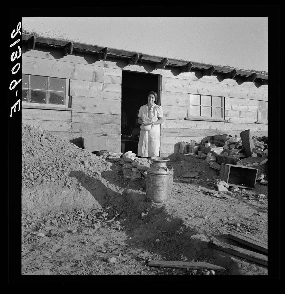The image is a black and white photograph depicting an individual standing in the doorway of what appears to be a modest wooden house with large windows. The person, presumably Mrs. Dougherty as indicated by the caption, stands confidently at the entrance wearing light-colored clothing that suggests a historical context or simple living conditions.

In front of the door lies a metal can on a concrete base, possibly used for storing liquids such as milk. To her right, there's an assortment of discarded items including what looks like old tools and containers scattered in disarray against a pile of dirt and rocks, hinting at either ongoing construction or abandonment. The setting appears rural with open skies above.

The structure itself is utilitarian with visible wood panels and lack of modern amenities such as electricity, suggesting it could be from an earlier time period when electrification was not prevalent, possibly the early to mid-20th century in a remote area like Warm Springs in Malheur County, Oregon. The photograph captures a moment that conveys simplicity, self-reliance or hardship associated with rural living during those times.