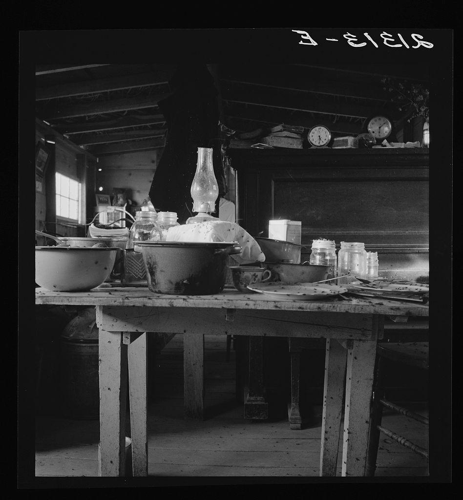 The image is a black and white photograph showing the interior of what appears to be an old-fashioned kitchen or dining area. On a large, wooden table with several legs can be seen various items typically found in such settings: two bowls, one filled with water, another empty; a bottle possibly containing oil, standing upright on its side; jars likely used for storage; and piles of stacked plates suggesting meal preparation or serving activities have taken place here. In the background, there is an individual whose identity remains unseen as they are only partially visible from behind, wearing dark clothing which could indicate that this person may be working in a kitchen environment.

The setting seems rustic with wooden beams supporting the ceiling overhead and various objects scattered around on shelves, including clocks, plates stacked upon each other, possibly for drying after use. The text "E-I-E-I-O" appears at the top right corner of the photo which is likely German phonetic notation for 'Ach! Ach!' indicating surprise or exclamation.

There's a sense of nostalgia and historical context in this image due to its monochromatic color scheme, suggesting it was taken during an earlier era. The presence of what seems like traditional kitchenware signifies that these scenes are from daily life activities within the household setting mentioned in the provided caption abou [...]