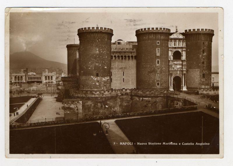 The image depicts a historical black and white photograph showing an old fortress with three prominent towers. These towers are cylindrical in shape, made of stone or brick, featuring battlements at the top for defense purposes. The central tower is more decorative with intricate architectural details including a statue on its peak.

In front of these towers lies what appears to be a large open space possibly used as a courtyard or parade ground. A wide staircase descends from this area leading down into another section that seems darker, suggesting it could lead underground or beneath the fortress walls.

The background features an urban landscape with buildings and structures indicative of late 19th-century architecture. There is also visible text on the photograph which reads "3 - NAPOLI - Nuova Stazione Marittima e Castello Angioino," identifying this as a new maritime station in Naples, Italy, along with the name 'Castello Angioino'. The setting appears to be historical and has an air of antiquity.

The photo's sepia tone adds to its vintage feel. There are visible marks on the photograph such as creases, stains, or tears suggesting it is quite old. This gives a sense that the image captures not only a moment in time but also carries with it history from Naples' past.