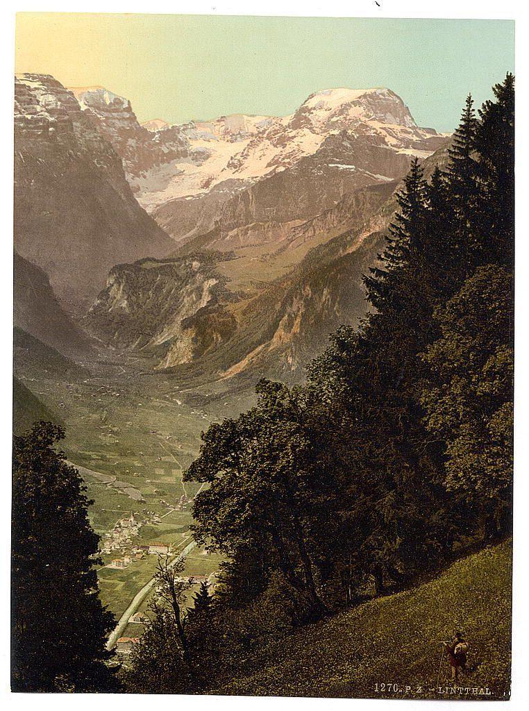 The image depicts a picturesque landscape featuring snow-capped mountains, lush green valleys, and dense forests. The scene captures the natural beauty of Switzerland with clear skies above where sunlight gently illuminates parts of the terrain. In the foreground, on the right side, there's a lone figure standing in silhouette against the verdant hillside, suggesting a sense of scale to the vastness around them. This person appears small compared to the towering peaks and expansive valleys that dominate the landscape.

The middle ground reveals rolling hills with patches of snow visible at higher elevations. Below these slopes is a river snaking through the valley, its path leading towards what seems like human habitation in the form of buildings nestled among fields. The overall composition of this image evokes tranquility and grandeur, capturing an idyllic snapshot of Switzerland's natural splendor during late spring or early summer.

There are textual elements present within the photo: "1270 P.Z LINTHAL" is inscribed at the bottom right corner in a script font that may indicate either a location or perhaps a date and reference related to this photographic print. Additionally, there's some text on top of the image which could be part of an album title or description but it’s not entirely legible due to its partial cropping out from view.

This photograph is likely histo [...]