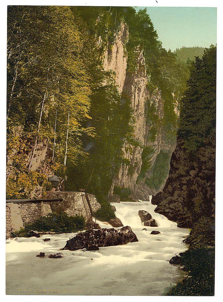 The image depicts a scenic natural landscape featuring rapid, turbulent water flowing through the middle of the picture. The river is flanked by rocky cliffs and lush greenery on either side, indicating a possibly forested area or park-like setting. Trees with bright yellow leaves suggest it might be late summer or early autumn. There are large boulders in the stream that disrupt its flow. In the background, dense foliage rises against steep rock faces, hinting at an elevated elevation. A stone wall appears to run alongside a pathway or road on the left side of the riverbed, suggesting human development near this natural wonder. The overall atmosphere is one of wild beauty and untouched nature, possibly a popular site for hiking or sightseeing given its prominent display in photochrom prints.