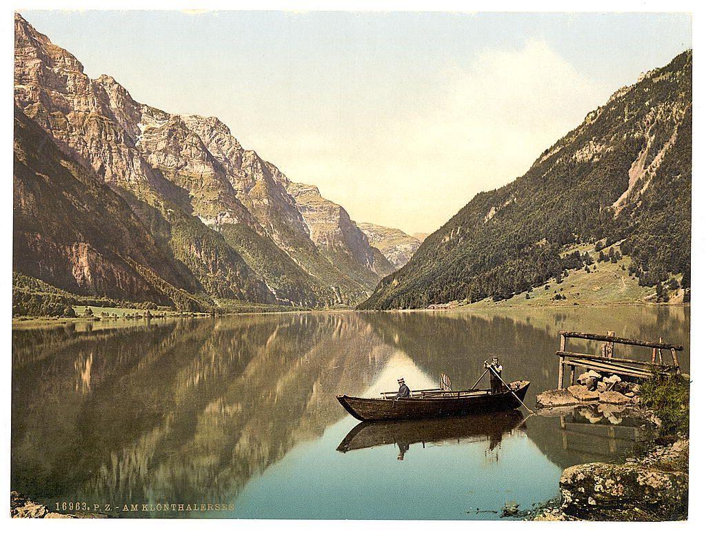 The image depicts a serene lakeside scene with stunning mountainous backdrops. The calm lake mirrors the rugged cliffs and greenery of the surrounding landscape, creating reflections that enhance its tranquility. A wooden rowboat is moored near a rustic dock on one side of the lake, where two individuals appear to be engaging in a leisurely activity such as fishing or boating. One person sits at the stern while another leans forward with an oar poised for action, suggesting they are navigating through calm waters.

The mountains rise steeply from the lakeside and are adorned with patches of greenery indicative of vegetation clinging to their rocky surfaces. The foliage is sparse enough to reveal glimpses of the cliff faces beneath, displaying a mix of earthy tones ranging from light brown to dark grayish hues. Some areas show traces of white snow or frost, hinting at varying altitudes and temperatures within this picturesque setting.

The sky above has a soft gradient transitioning from pale yellow near the horizon line where it meets the mountains, gradually fading into lighter blue as one looks upward towards what appears to be a partly cloudy day. The overall atmosphere is that of an idyllic natural retreat with historical undertones suggested by its vintage look and mention of Klonthal Lake in Switzerland during approximately 1890-1900.

Additional information about t [...]