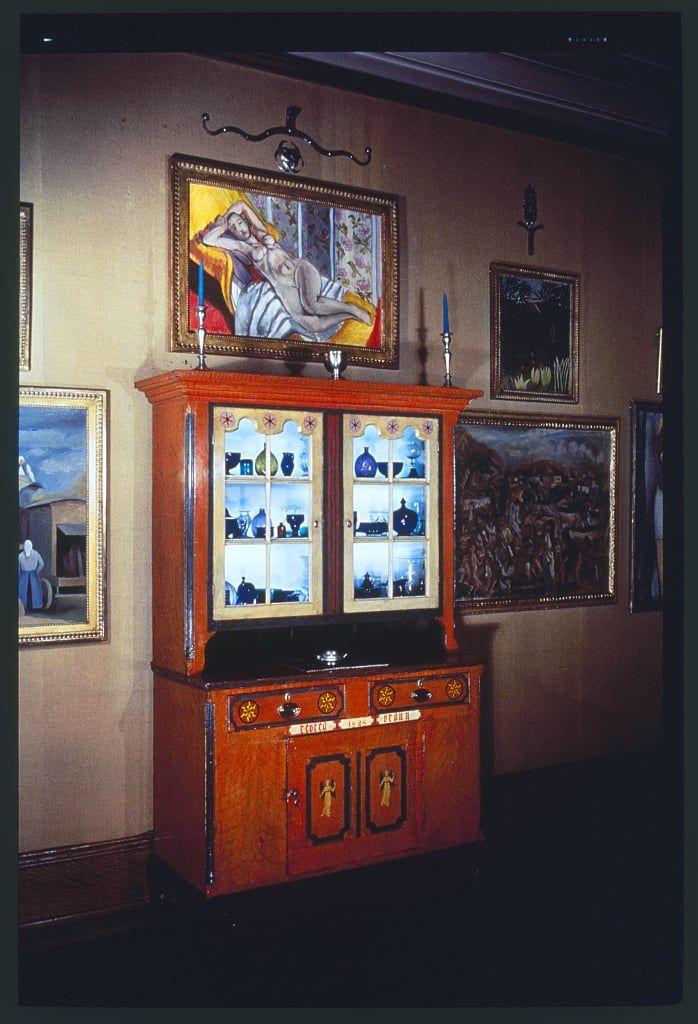 The image depicts an interior room with a collection of framed artworks displayed on the walls and housed within decorative cabinets. The central focus is a wooden cabinet, rich in red-brown tones, featuring ornate brass detailing and a glass front showcasing an array of blue bottles or vases arranged neatly in rows.

Above this cabinet hangs another prominent artwork depicting two reclining figures with dynamic color contrasts against a warm background. To the left and right are smaller paintings; one shows a figure standing beside a classical urn, while others include abstract patterns and possibly still-life compositions featuring flowers.

The room's ambiance is enhanced by subtle lighting that casts gentle shadows on the walls, creating an intimate atmosphere suitable for appreciating art collections. The overall impression suggests this setting could be part of an exhibition or within a private collection curated with discernment in mind.
