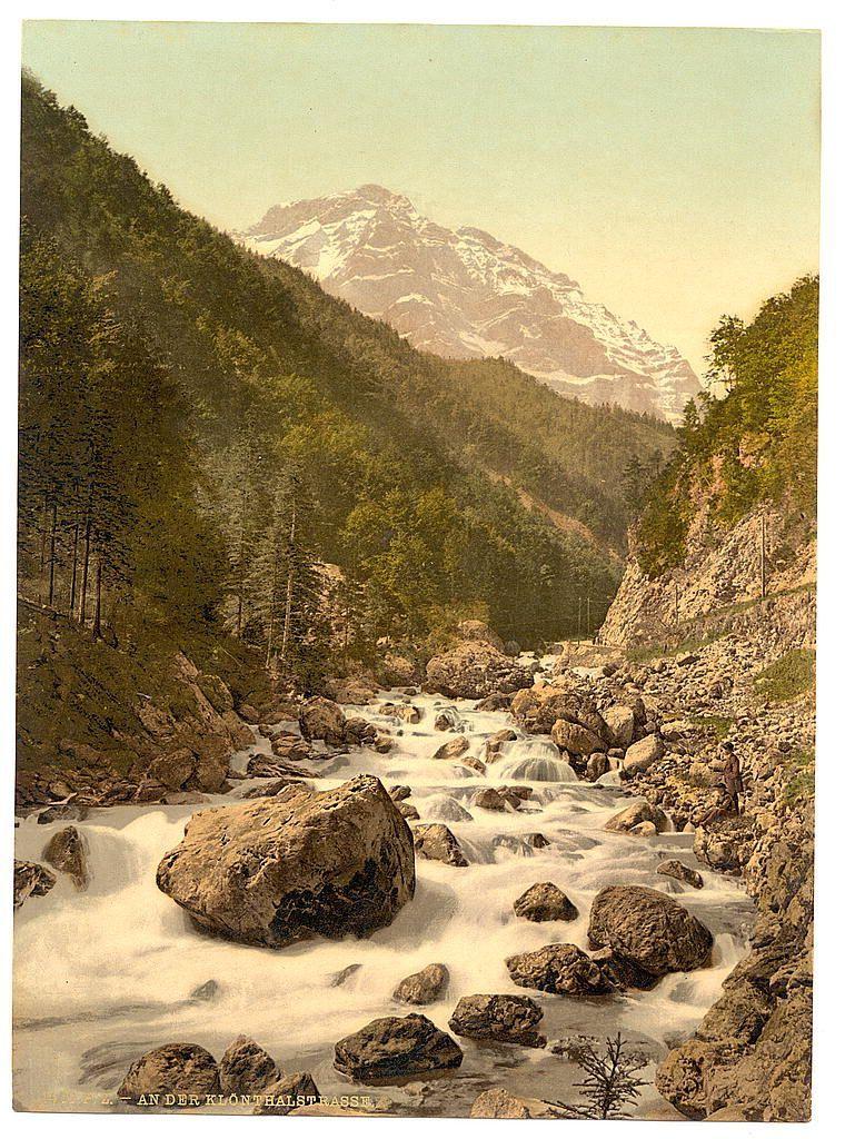 The image depicts a picturesque mountain landscape with flowing water and rocky terrain. A river or stream meanders through the scene, surrounded by large boulders that create small rapids as they rush over them. The surrounding environment includes dense green forests on both sides of the watercourse, leading up to towering mountains in the background which are adorned with patches of snow at higher altitudes indicating their peak elevation is quite high.

The overall color palette has a warm, vintage tone suggesting this could be an old print or postcard possibly dating back around the late 19th century. The naturalistic scenery suggests that it was captured during daylight hours under clear skies. Notable features include the detailed rendering of tree textures and mountain snowcaps which gives depth to the image.

There is a small human figure visible near one of the boulders, providing a sense of scale for the vastness of nature surrounding them. This indicates humans are present but not overtaking or altering the natural landscape significantly at this moment in time captured by the photograph.