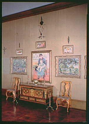 The image shows an elegant room with a vintage aesthetic, featuring classic artwork displayed on the walls. There are four framed paintings hung symmetrically above a decorative table in the center of the composition. The pieces include impressionist works depicting various landscapes and possibly scenes from everyday life.
Two antique chairs flank the painting arrangement, adding to the old-world charm of the setting. A wooden cabinet with intricate carvings sits below the tableau, further emphasizing the room's historic feel. The flooring appears polished wood, enhancing the overall warm ambiance within this refined space.
This specific image is part of Dr. Albert Coombs Barnes' collection at the Barnes Foundation in Philadelphia and dates back to August 1942 according to the caption provided by Gottschoschleisner Inc., a company known for its archive of museum images, including photographs from various institutions such as the Museum of Modern Art.
The image itself measures approximately five inches wide by seven inches tall on transparency.
