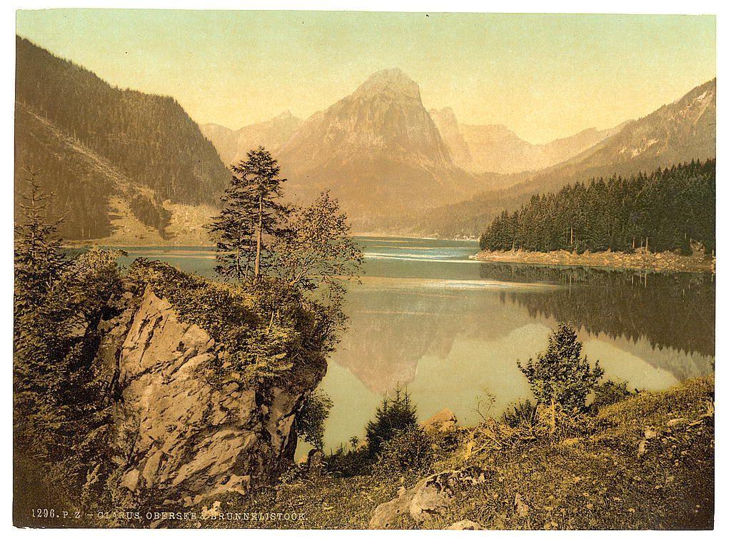 The image depicts a serene landscape featuring a tranquil lake nestled within mountainous terrain. In the foreground, there's an imposing rock formation on the left with sparse vegetation and several trees scattered around it. The midground is dominated by calm waters reflecting the surrounding environment, including coniferous forests lining its edge. Rising majestically in the background are towering mountains shrouded partially in mist, suggesting either early morning or late afternoon lighting conditions. There's a sense of untouched wilderness with no visible human structures, evoking a timeless natural beauty.