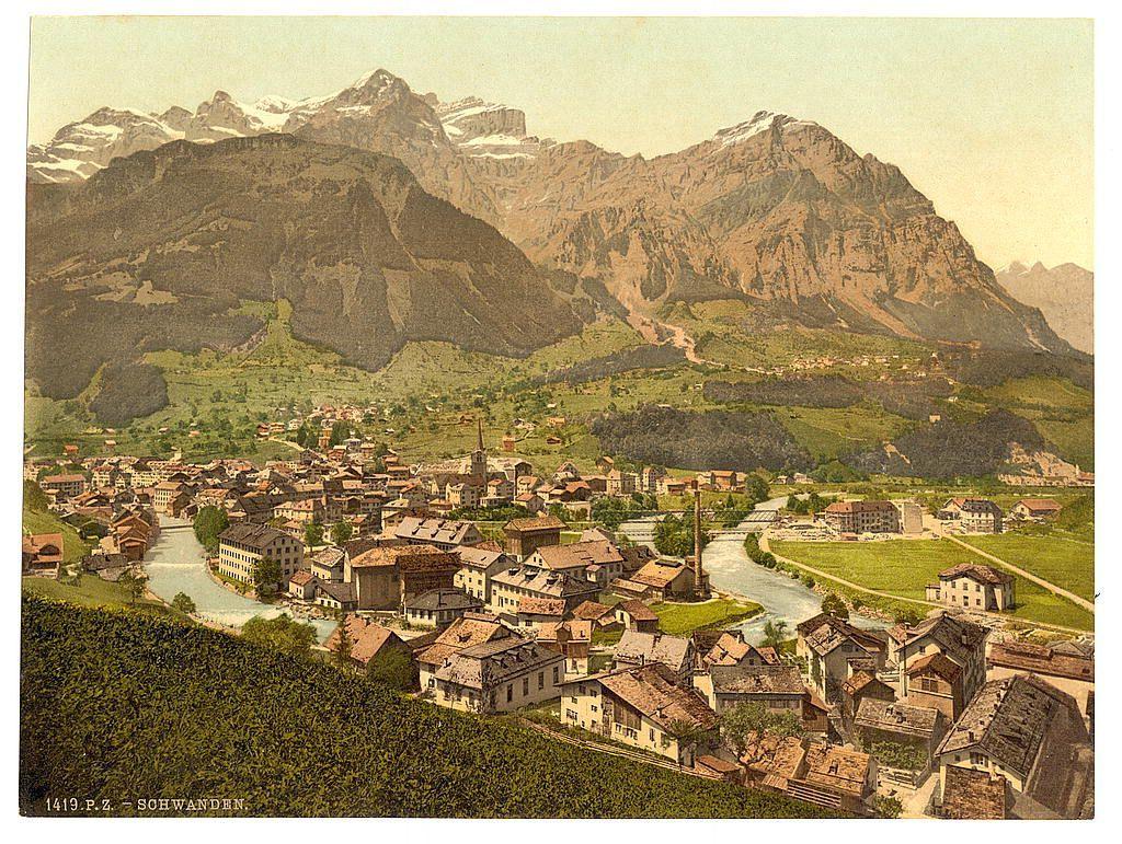The image presents a picturesque townscape nestled amidst the backdrop of imposing mountains. The foreground features lush greenery, transitioning into terraced fields that cascade down to an intricate network of houses and buildings with steeply pitched roofs, typical of European alpine architecture. A river meanders through the town, reflecting sunlight on its calm waters, which is bordered by pathways lined with vibrant wildflowers.

The central part of the image showcases densely packed residential structures, their earthy tones complementing the natural landscape around them. Among these buildings are churches and other public edifices, distinguishable by their steepled roofs and bell towers. The architecture exhibits a variety of styles but maintains a cohesive aesthetic that harmonizes with its surroundings.

In contrast to this human settlement, large green fields dominate the lower part of the image, likely dedicated to agriculture or pastureland for livestock. This open space provides a stark yet pleasing juxtaposition against the tightly packed town and serves as an invitation into the tranquil life in Switzerland's alpine regions.

The mountains rise majestically behind the town, their rugged peaks adorned with patches of snow, hinting at higher altitudes not far from this quaint settlement. The sky above is rendered in muted hues, suggesting a clear day or po [...]