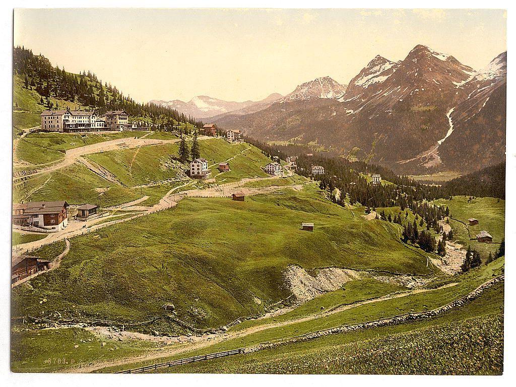 This image depicts a picturesque Alpine landscape featuring rolling green hills and majestic mountains in the background. There are several chalet-style buildings scattered across the hillside, with some situated on terraced areas to adapt to the slope of the terrain. The architecture is typical of mountain resorts or rural settlements found in alpine regions. Snow-capped peaks suggest that this location experiences a cold climate, possibly during late spring, early summer, or autumn when snow has not fully melted but still persists at higher elevations.

The foreground shows lush green fields with traces of human activity such as paths and small fences. The overall color palette is dominated by greens from the vegetation, contrasted against the earthy tones of the buildings and rugged mountain terrain. This image likely represents a historical view given its vintage style, indicating it could be an early photograph or postcard capturing the beauty and serene atmosphere of Switzerland's mountains during that era.

The specific mention "Arosa" in the reference suggests this location might have been Arosa, Grisons, Switzerland. Between ca. 1890 and ca. 1900 is a timeframe when such picturesque Alpine scenes were popular for tourism promotion or personal mementos of travel. The image source credits Loener.nl as providing information about the photochrom print with reference  [...]