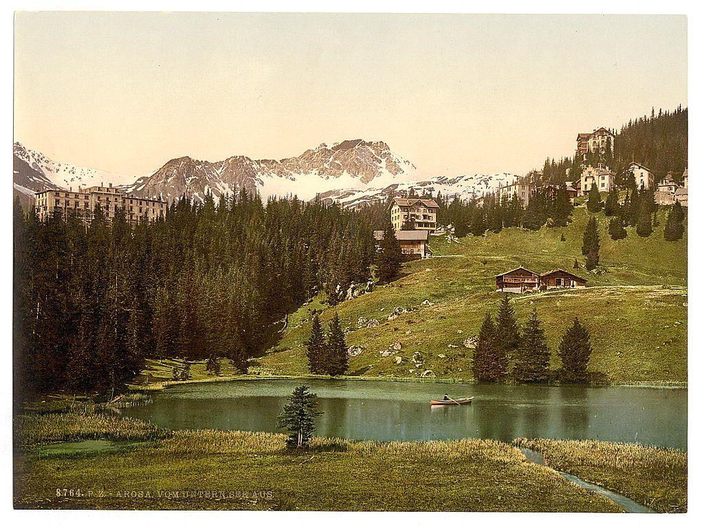The image is a vintage postcard depicting an alpine landscape, likely from the late 19th or early 20th century. It shows a serene lake nestled in a valley surrounded by rolling hills and lush greenery. In the background, majestic mountains with snow-capped peaks rise against a pale sky. The foreground features tall evergreen trees bordering the water's edge.

On either side of the pond are rustic cabins or chalets, indicative of early mountain lodges or homes. A small wooden bridge spans across the lake in one area, and there is visible human activity with at least two boats on the waters, suggesting recreational use such as boating for leisure. The coloration suggests it's a chromolithograph print due to its muted yet naturalistic hues.

The text "8764 P.Z - ARORA VOM UNTERN SEE AUS" appears in the bottom left corner of the image. This likely identifies the location or publisher associated with this particular postcard series, and references 'Arosa' as a notable Swiss town within Grisons (Graubünden) canton in Switzerland.

This scene exudes tranquility and offers insight into how such picturesque alpine regions were perceived and marketed during that era.