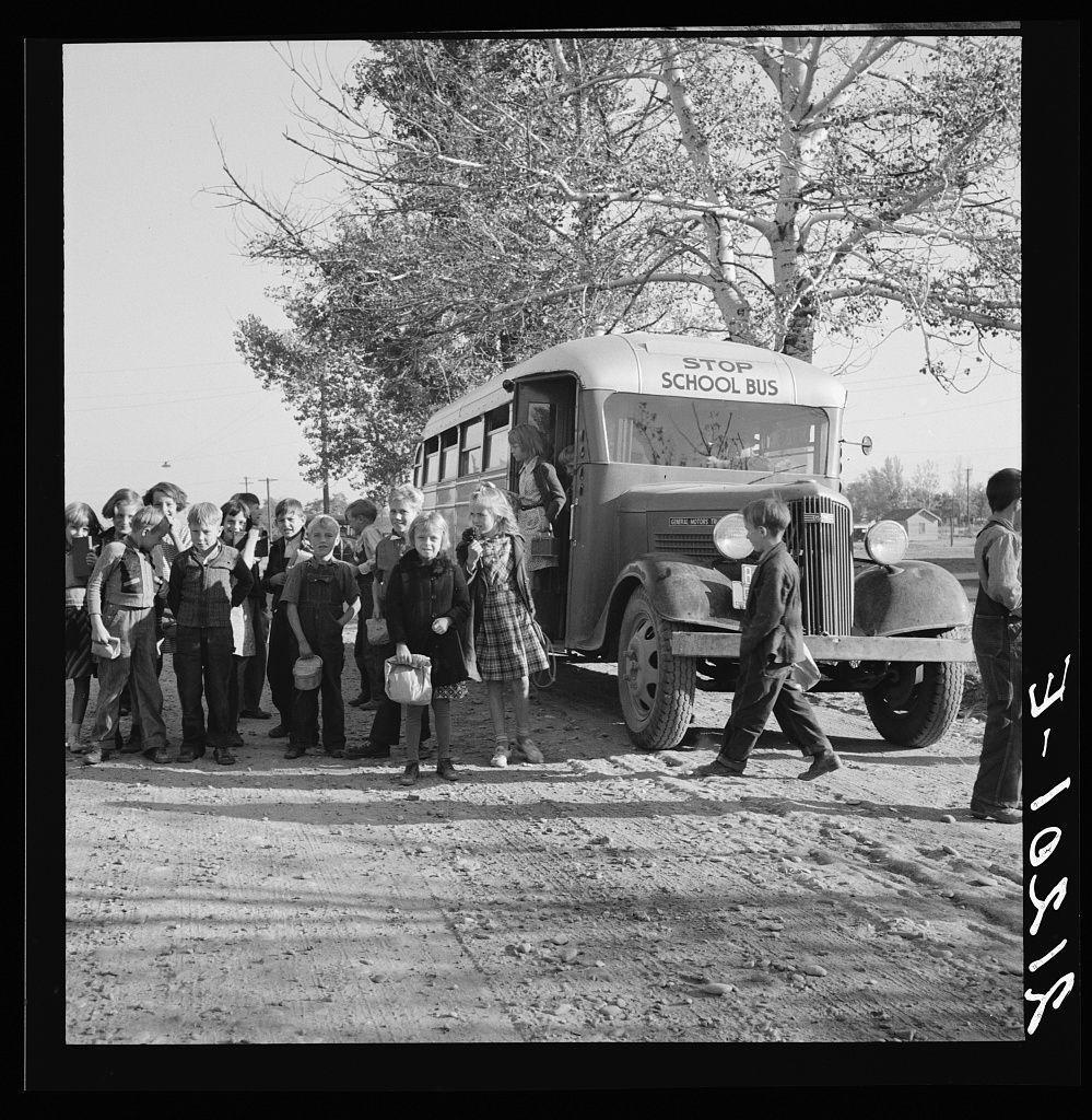 The image is a black and white photograph depicting several children gathered beside an older model school bus. The setting appears to be rural, with barren trees in the background suggesting it could be late fall or winter. Most of the children are standing on what looks like unpaved ground with patches of grass. A few individuals can also be seen seated by a tree.

The expressions and body language of the kids vary; some appear joyful while others look more reserved. The bus is marked "School Bus" along its side, indicating it's used for transporting students to school or other educational activities. There are no visible signs providing information about specific locations within Oregon.

Additional context can be derived from a caption noting that this photo was taken in Ontario and Malheur County, suggesting the area could have historical significance regarding education during times when such rural bus services were common practice. The overall mood is nostalgic, reflecting on simpler times of schooling for children who lived outside urban centers.