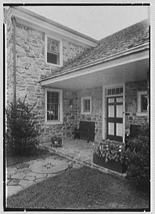 The image is a black and white photograph of an old stone house with a covered porch. The entrance has double doors framed by two small, square windows above them. To the right side of the entrance are steps leading up to another door that opens into what appears to be a sunroom or enclosed patio area with glass sliding doors.

The porch is supported by wooden beams and columns painted in dark colors, contrasting against the stone facade of the house. The ground around the porch features irregularly shaped flagstone paving stones laid out haphazardly.

There are two potted plants placed on either side of the entrance steps, adding a touch of greenery to the scene. On the left side of the image is part of what looks like a small garden or lawn area with grass and possibly some shrubs.

The overall impression is one of a quaint, rustic home that has been preserved over time with care for its historical architecture. The photo seems to capture an entrance detail from August 20th, as mentioned in Dr. A.C. Barnes's record about the residence located in Chester Springs, Pennsylvania known as "Ker-Feal."