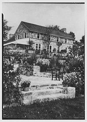 The image depicts a large, two-story stone house with multiple windows and an overhanging roof. The house is surrounded by lush gardens filled with various plants, flowers, and shrubs. A wooden bench sits in the foreground of the garden area, inviting visitors to sit and enjoy the view.
The photograph appears to be black-and-white, suggesting it may have been taken several decades ago. There are two chairs visible on a porch or terrace that extends from the house, adding an element of comfort and relaxation to the scene.
Overall, this image conveys a sense of tranquility and picturesque beauty in a suburban setting during the mid-20th century.