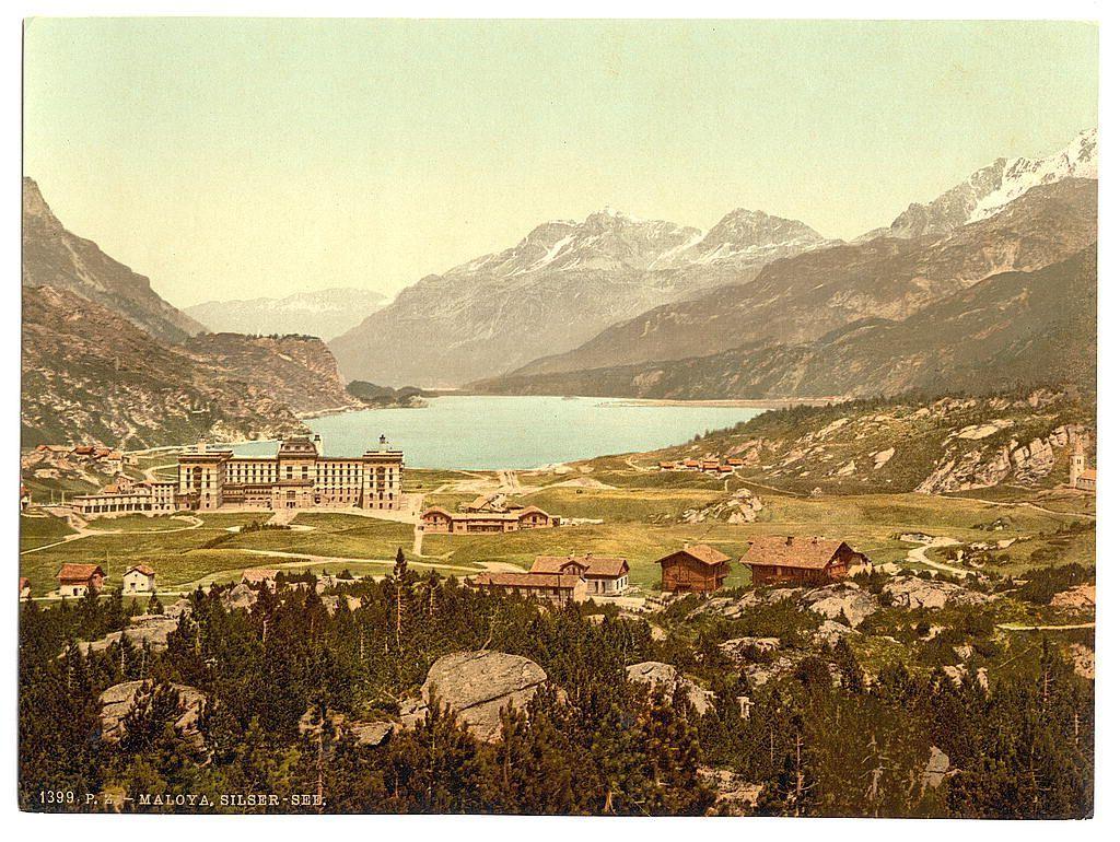 The image depicts a picturesque landscape with rolling mountains, dense forests, and a serene lake nestled within the valley. In the foreground, there are traditional wooden chalet-style buildings scattered across a grassy field, indicative of alpine architecture. The middle ground is dominated by an expansive building complex on the lakeside, featuring classical architectural elements such as large windows and symmetrical design, which suggests it could be a grand hotel or spa resort. In the background, majestic mountains rise with patches of snow still clinging to their peaks, hinting at the region's high altitude and colder climate.

The overall scene conveys an early 20th-century European holiday destination, capturing both natural beauty and human-made elegance harmoniously coexisting in a mountainous setting. The coloration has faded hues typical of historical photographs from that era, giving it an antique look reminiscent of postcards or old travel catalogues showcasing Swiss tourist spots during the late Victorian period.