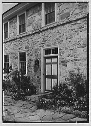 This black and white photograph shows a two-story stone house with several windows visible on both floors. The entrance is via the rear of the building, as indicated by an exterior door that opens onto what appears to be a small patio or landing area made from cobblestones. Surrounding the doorway are bushes and flowers planted in beds along the foundation walls. A mailbox can be seen attached to one of the stone walls near the top floor windows. The overall condition of the house suggests it is well-maintained, with no visible damage or signs of neglect.