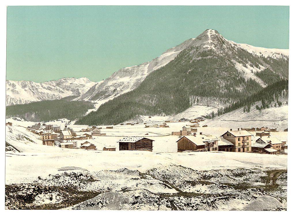 The image depicts a picturesque winter scene set in the Swiss Alps, specifically around Davos. Snow blankets the ground and rooftops of numerous wooden chalet-style buildings spread across a valley floor, indicating an old mining town or settlement with traces of industrial activity from earlier times. The architecture is simple yet functional, featuring sloped roofs designed to shed snowfall effectively.

In the background stands a prominent mountain peak dusted lightly with snow, its slopes dotted sporadically with trees suggesting sparse vegetation adapted to high-altitude conditions. Above it looms another larger range, providing depth and scale to the composition. The sky is clear with no visible clouds, hinting at calm weather typical of this region.

The image appears aged, rendered in sepia tones rather than full color, which conveys a sense of nostalgia or historical documentation. This specific photograph seems part of a collection from "Views of Switzerland" by Loener's Photochrom prints series, dated approximately between 1890 and 1900, suggesting it captures the region during its late Victorian period.

Overall, this image encapsulates the serene beauty and stark rural life of an early twentieth-century Swiss winter landscape.