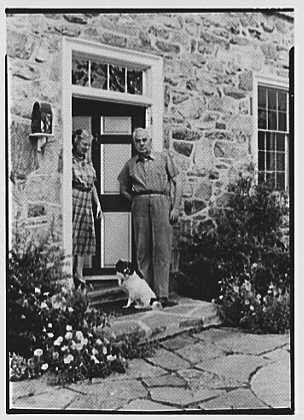 This black and white photograph shows an older man and woman standing on the front porch of a stone house. The man is wearing glasses, suspenders, and a long-sleeve shirt with rolled-up sleeves, while the woman has her arms crossed over her chest. They are both smiling as they look towards the camera.

A small dog stands in between them, looking directly at the camera with its tongue sticking out. There's also another small black-and-white cat standing on top of a low wall near the front door. The house features brick and stone construction, with windows that have glass panes featuring geometric designs. On either side of the doorway are built-in mailboxes.

In the foreground, there is a garden filled with blooming flowers. Some shrubs can be seen in the background as well. This image appears to capture a moment from daily life during August 20th,1942 at Dr. A.C. Barnes' residence located on Ker-Fell Street in Chester Springs, Pennsylvania.

The photo is credited to Gottscho-Schleisner Inc., which was likely the photography studio responsible for taking this photograph. The image appears aged and has a vintage feel due to its black-and-white composition as well as faded colors around certain areas like flowers or shrubs indicating age-related wear on photographic materials over time.

Overall, it's an adorable snapshot of two individuals enjoying their porch with pets ami [...]