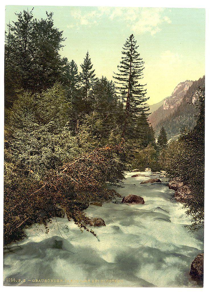 This image depicts a serene winter landscape, featuring a river with its banks partially covered in snow and ice. The water appears clear and moves gently over rocks that protrude from the surface. On either side of the stream, lush greenery is visible, including various trees like conifers and deciduous varieties, suggesting this scene could be captured during late autumn or early spring when temperatures allow for both seasonal foliage and snowy ground. In the background, towering mountains with rocky outcrops are partially shrouded in mist, adding a sense of depth to the composition. The color palette is dominated by cool blues and greens from the trees and water, contrasted against the muted browns of the tree trunks and snow-covered landscape. This picturesque scene likely evokes feelings of tranquility and solitude associated with natural winter settings.