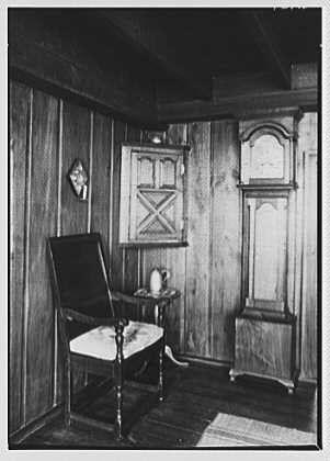 This black and white photograph depicts an interior room with wooden walls, floors, and a single dark chair. The atmosphere is rustic, as the wood shows signs of aging or neglect. A framed picture hangs on one wall above what appears to be a small sideboard table holding various items including a jug and some other miscellaneous objects. On another wall sits a standalone cupboard with shelves that have an intricate design at its center. In front of this cupboard is a wooden bench, which has no backrest or arm rests, suggesting simplicity in the room's furnishings. There are also what seem like papers on the floor near the chair, indicating disarray within the space. A rug partially covers part of the hardwood flooring and adds some texture to the scene. Overall, there seems to be a story behind this setting which could potentially hint at historical or personal significance linked with Dr. Barnes' residence in Chester Springs, Pennsylvania as suggested by the caption provided.