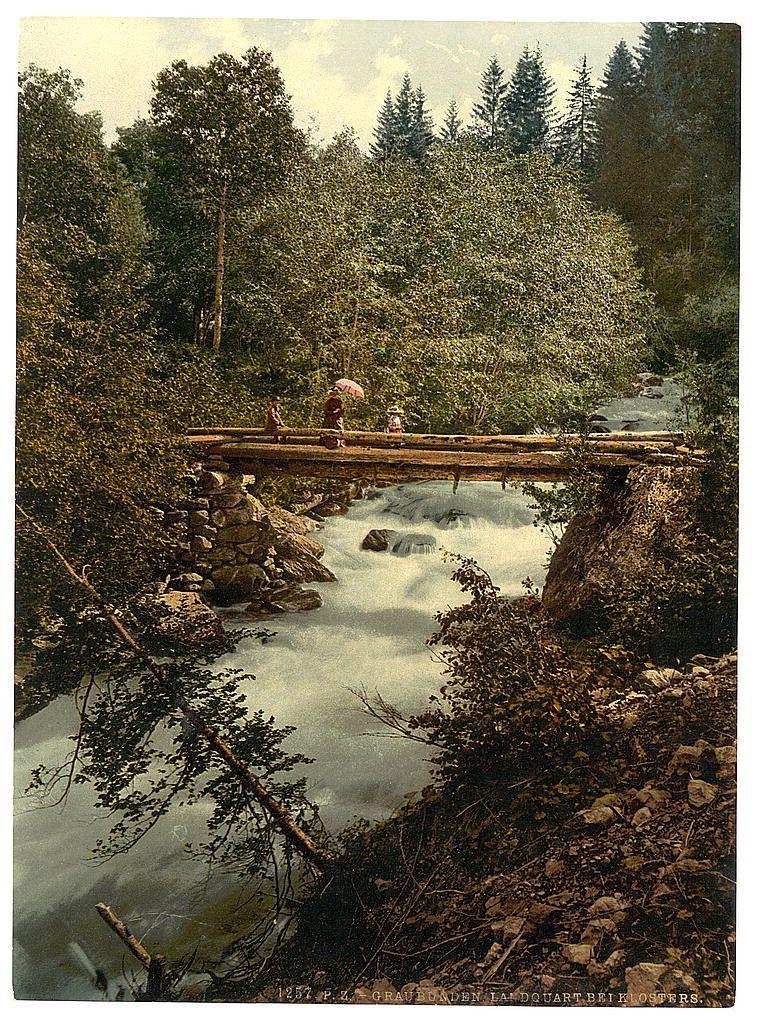 The image depicts a serene natural scene featuring a wooden footbridge spanning over a gently flowing river. The bridge is constructed from logs and branches, with some sections appearing more weathered than others. Two individuals are seen crossing the bridge; one holds an umbrella while navigating its length.

Lush greenery surrounds the area, including trees in various shades of green and tall evergreens that frame the background. Sunlight filters through the foliage, casting dappled shadows on the river's surface below. Rocks line parts of the riverbank, adding texture to the scene.

The overall atmosphere is tranquil, suggesting a leisurely stroll or perhaps an excursion into nature during what appears to be late spring or summer. The image has the distinctive appearance of early 20th-century colorized photography, with its rich hues and somewhat muted tones that emphasize warmth over vibrancy.