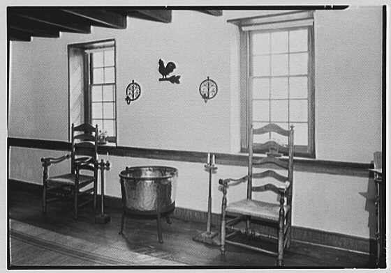 The image depicts a black and white photograph of an interior space, possibly from the early to mid-20th century. It shows three wooden chairs arranged around what appears to be a large metal cauldron or pot placed on a low table in the center of the room.

On the wall behind the cauldron are two decorative rooster figures mounted above small round mirrors that serve as clock faces, suggesting they might indicate time rather than actual clocks. The walls have horizontal paneling with visible joints and nails holding it together, indicative of traditional construction methods.

The floor seems to be wooden planks laid parallel to each other, extending from the foreground into a distance where no furniture or objects are present. Two windows allow natural light into the room; one has bars across part of its lower half while the second appears fully intact and unlatched.

The overall atmosphere evokes simplicity and modesty with an emphasis on functionality over ornamentation. The absence of modern elements like electronic devices, clocks displaying accurate times, or decorative objects such as paintings indicates this scene could be from a historical setting.