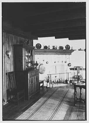 The image shows a black and white photograph of an interior space, likely taken in the early 20th century. The room appears to be a dining area with wooden furniture including chairs, tables, and a china cabinet filled with various dishes and glassware on display shelves above it.
In the background, there is a large window that lets natural light into the room, showing some outdoor scenery outside of the building's roofline. A rug can also be seen on the floor near one of the windows.
The overall atmosphere suggests an old-fashioned or vintage aesthetic with a focus on practicality and utility in design. The absence of color adds to this timeless feel as it emphasizes textures such as wood grain, glass reflections, and fabric patterns.