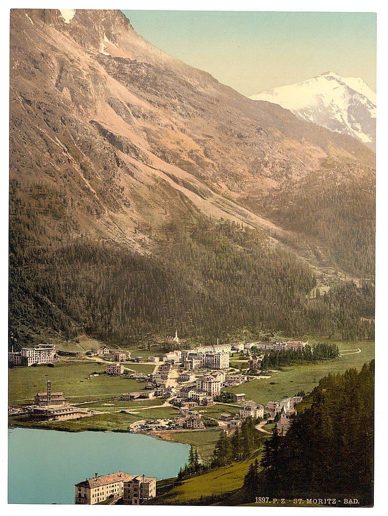 The image is a vintage postcard depicting the picturesque town of St. Moritz in Switzerland during late 19th or early 20th century times, as indicated by its caption "St-Moritz - Bad," dating back to around 1397 P.Z.. The village sprawls across a valley floor surrounded by towering mountains and lush greenery with coniferous trees. Prominent buildings are spread throughout the scene, including what appears to be luxury hotels or resorts with distinct architectural styles reflecting European design sensibilities of that era.

In the foreground lies a body of water—a lake—reflecting some structures on its shorelines. The color palette is rich and warm, featuring greens from the vegetation, blues in the water bodies, and earth tones for the buildings and surrounding landscape. Notable natural features include Piz Surlej's snow-capped peak looming over the settlement.

The postcard showcases a panoramic view that likely aims to convey both the charm of St. Moritz as an alpine retreat and its status as a well-known tourist destination during its time, potentially highlighting its appeal for leisure travel given the mention "St-Moritz - Bad," which translates from German to mean spa town or resort.

Further information about this specific image can be found on Views of Switzerland website at https://images.loener.nl/PhotochromPrints/full/65e4/65e47a255890ae069f67c182.jpg.