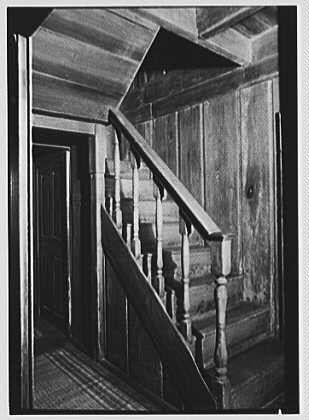A black and white photograph of a wooden staircase with railings leading up to an upper floor. The image shows the stairs winding around the turn, supported by turned balusters on each side. A door is visible in the background at the base of the stairwell. The style of construction suggests it may be from the early 20th century or earlier, indicative of a historical residence.