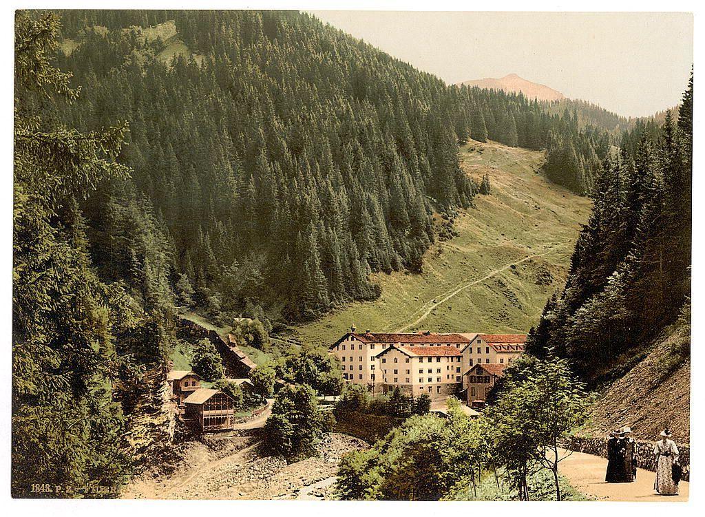 The image depicts a lush, forested mountain landscape with dense coniferous trees covering rolling hills. In the foreground and middle ground, there are several buildings in an alpine architectural style, suggesting this could be a small village or resort nestled within nature. The coloration of the photograph is sepia-toned, typical for images from that era, indicating it may date back to the late 19th century.

In the bottom right corner, two individuals appear to be walking along a dirt path; their attire suggests historical fashion consistent with the Victorian period or possibly early Edwardian times. One person appears to wear dark clothing and carries an umbrella while the other wears lighter-colored dress with what seems like a hat, both dressed in styles common for women during that time.

The image is marked "1843" at its bottom left corner which could be related to either the date of the photograph or perhaps part of a series. The overall scene evokes an impression of leisure and retreat into nature, characteristic of spa resorts popularized by European tourism in the late 19th century.

Further information about this image can be found on Views of Switzerland website with reference number Pratigau, the Baths of Fideris, Grisons, Switzerland between ca. 1890 and ca. 1900: https://images.loener.nl/PhotochromPrints/full/65e4/65e47a255890ae069f67c186.jpg