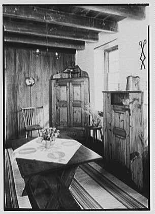 A black and white photograph of an antique dining room, featuring a wooden table with a floral centerpiece. The furniture includes benches on either side of the table and chairs at one end. In the background is a large armoire-style cupboard beside which stands another cabinet or chest. Decorative elements such as wall sconces add to the vintage ambiance. This image appears to be from Dr A.C. Barnes' residence in Chester Springs, Pennsylvania, dating back to August 20th of an unspecified year. The photo was captured by Gottschol-Schleisner Inc., and is stored at Loener NL under their collection number "691e/691efa90450e57a2674fc528.jpg".
