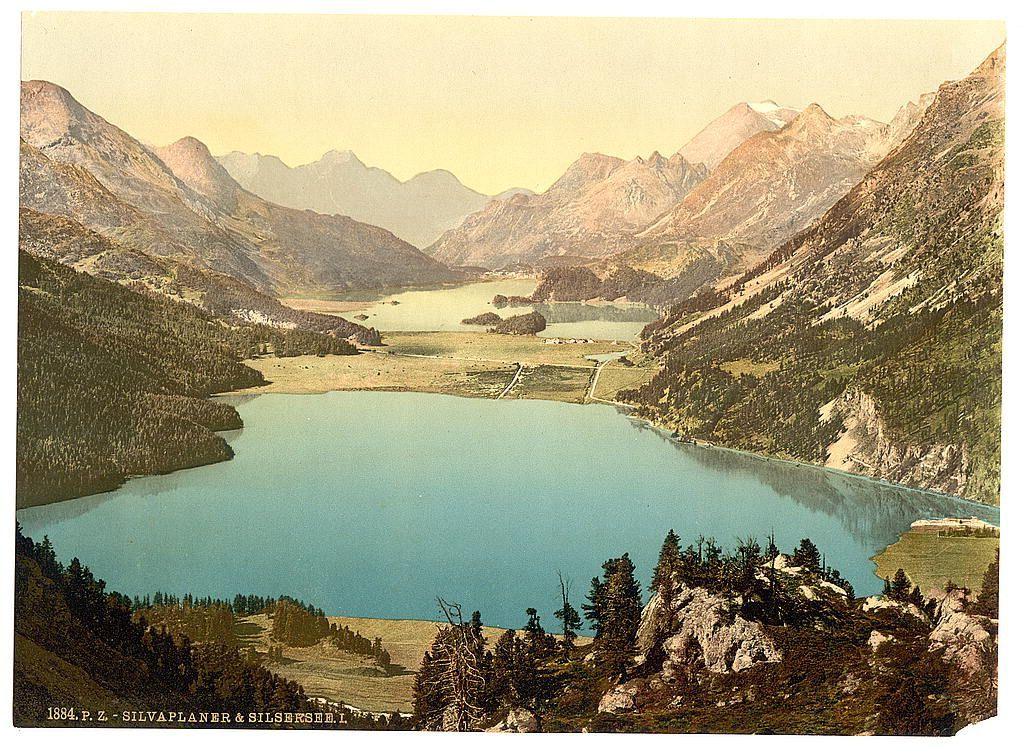 This image depicts a scenic landscape featuring a large, tranquil lake nestled amidst mountainous terrain. The foreground is dominated by lush greenery and clusters of trees that transition into rocky outcrops near the water's edge. A prominent cliff adorned with sparse vegetation extends from the left to right across the mid-ground, leading down to the expansive blue waters below.

In the distance, majestic mountains rise dramatically against a pale sky, their slopes covered in various shades of green and brown, indicative of forested areas interspersed with exposed rocky surfaces. Nestled at the base or within the valleys between these towering peaks are patches that resemble settlements or clusters of buildings, though details are indistinct from this vantage point.

The coloration suggests a vintage postcard print, likely dating back to late 19th-century Europe. The presence of text stating "Silvaplana & Silselsee" and the year "1884 P.Z." supports its Swiss origin in Engadine region. This image encapsulates the serene beauty and picturesque allure typical of Alpine landscapes during that era.