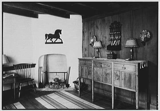 The image depicts an interior scene with a rustic and vintage atmosphere, possibly from the early 20th century. A wooden wall adorned with various decorative items such as a horse silhouette, patterned textiles or plaques, and a mounted clock set against it is visible on the right side of the frame.
In front of this textured backdrop lies an old-fashioned cabinet-style dresser, displaying three drawers above two doors that open to reveal storage compartments. Each drawer features ornate handles with intricate patterns. To its left stands what appears to be a heater or small stove covered in dust and cobwebs indicating age and disuse. Above the fireplace is a shelf housing more items, including books, baskets, and possibly other artifacts.
The room also contains wooden chairs, one placed near each side of the cabinet-dresser; both are empty with vertical slats on their backs. The floor seems to be covered by an area rug featuring stripes or patterns in varying shades. Overall, this scene captures a snapshot from another era showcasing antique furniture and décor within a homely setting.