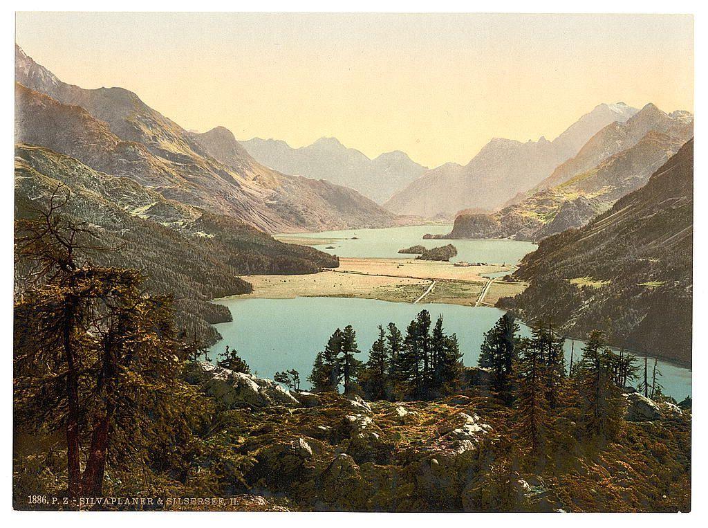The image depicts a serene, expansive view of a mountainous landscape with lush greenery and rocky outcrops. In the foreground, we see clusters of pine trees on rugged terrain covered in yellowish-brown foliage, indicative of either autumn or early winter conditions. The middle ground is dominated by a calm turquoise lake nestled amidst rolling hills, reflecting hints of light blue sky above. On one side of this body of water extends flat agricultural land with visible structures and pathways suggesting human habitation. In the background, majestic mountains rise steeply from the valley floor, their peaks partially cloaked in mist or low clouds, adding a sense of depth and grandeur to the scene.

The color palette is rich yet muted, with various shades of green for vegetation, earthy tones on rocky surfaces, and blues ranging from turquoise within the lake to deeper hues at its farthest reaches. The overall atmosphere evokes tranquility and natural beauty, while also hinting at a connection between human life and the untouched wilderness.

At the bottom left corner, there is text that reads "1890 P Z SILVAPLANER & SILSVERSE II", suggesting this image could be part of an album or collection featuring Swiss landscapes. The mention of specific places such as Upper Engadine, Silvaplana, and Grisons indicates a geographical focus on the region in Switzerland known for its pict [...]