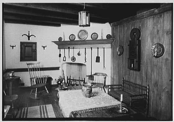 The image is a black and white photograph showing the interior of an old-fashioned room with wooden walls. It appears to be a dining area or kitchenette, featuring a table set for four people with chairs around it. The table has a decorative centerpiece on it.
On one wall above the fireplace, there are various objects such as plates, bowls, spoons, and ladles arranged in a line along the shelf. To the left of this arrangement is another similar display but smaller in size. There's also an empty chair with a spindle backrest near these displays.
The room has several decorative elements including wall sconces on either side of the fireplace area, circular plates mounted horizontally at various heights, and round mirrors or frames placed above them respectively. A wooden bench sits against another wall to one side while two additional chairs are positioned in front of it facing towards each other creating a conversation setting.
The overall atmosphere suggests an antique style with emphasis on simplicity yet rich detailing which was characteristic during early 20th-century American interiors, reflecting rural or colonial living conditions prevalent at that time.