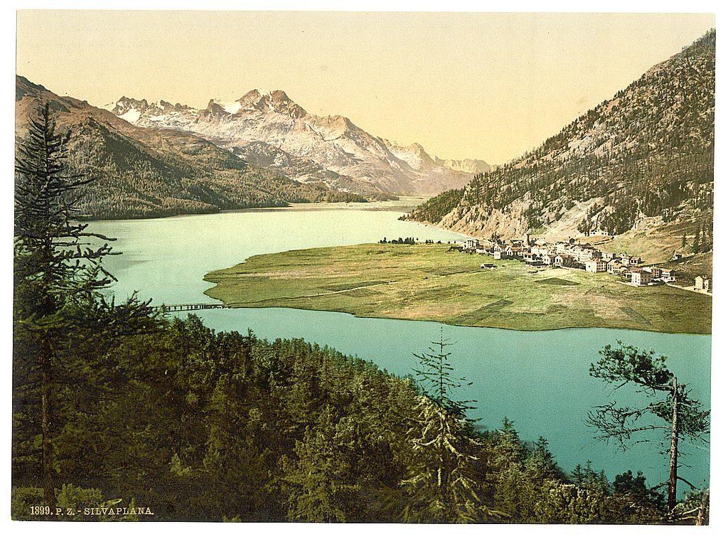 This image depicts a picturesque landscape featuring a serene lake nestled amidst lush green hills and towering mountains. The sky is clear, suggesting fair weather conditions. A quaint village with houses and buildings can be seen on the shore of the lake, providing a sense of human habitation within this natural setting. Tall trees are scattered throughout the scene, adding to the verdant atmosphere. The colors in the image have been printed in varying shades—greens for foliage, blues for water, browns for mountains—and there's an overall vintage look consistent with historical postcards or prints from that era.