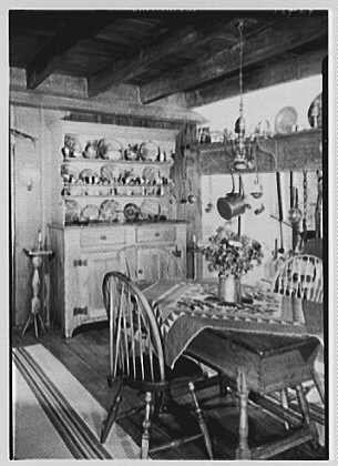 The image is a black and white photograph depicting an interior scene of what appears to be a dining room in a home. The setting has vintage furnishings, including wooden chairs around a table with visible wear on the surface indicating age or frequent use.

On one side of the photo stands a large cabinet adorned with various dishes displayed on open shelving above, showcasing different sizes and shapes of plates, bowls, and other dishware neatly arranged in rows. To its left is an end table supporting what seems to be books or decorative items placed vertically.

Hanging from the ceiling are two hanging lamps that cast light onto the dining area below. A vase filled with flowers sits on a surface beside one chair, adding a touch of natural beauty and color contrast against the monochromatic background.

The overall atmosphere conveyed by this image is nostalgic and reflects an earlier era's domestic life within American households during mid-20th century America.